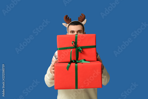Young man in reindeer horns with Christmas gift boxes on blue background
