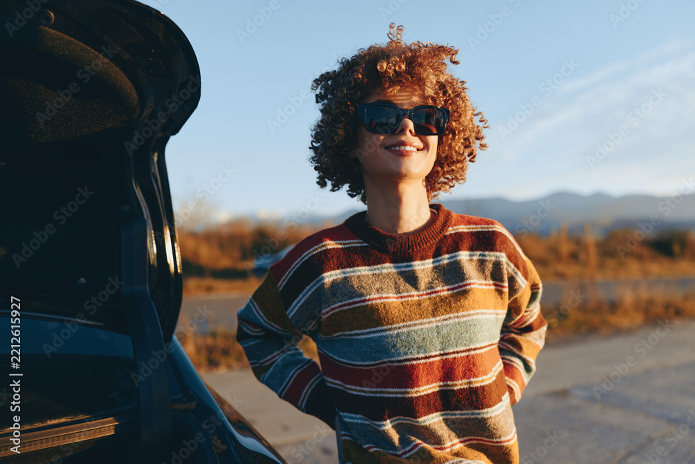 Obraz premium Woman with curly hair smiling outdoors, wearing striped sweater, standing near car on road, sunlight golden hour, relaxed lifestyle mood, soft breeze, warm natural colors, distant hills