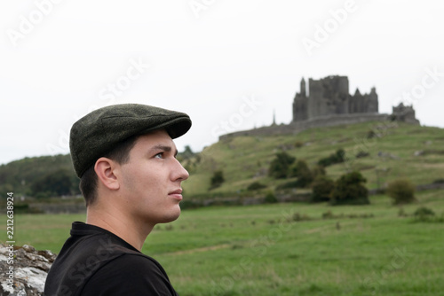 Young man wearing flat cap gazes toward a distant hilltop castle across rolling green fields, suggesting contemplation, travel and historic vintage atmosphere