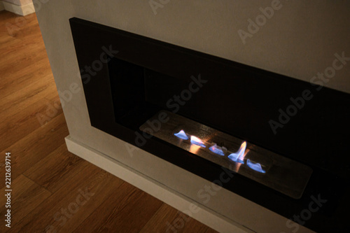 Sleek built-in bioethanol fireplace features matte-black frame, linear burner with dancing flames, seamless wall integration, warm glow contrasting light wood floor