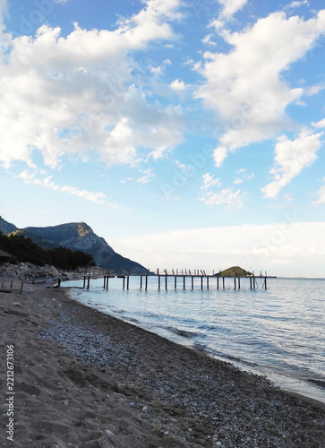 Scenic view of a pebble beach and calm blue sea bay with green forested rocky cape under blue sky with clouds, Mediterranean coastal landscape