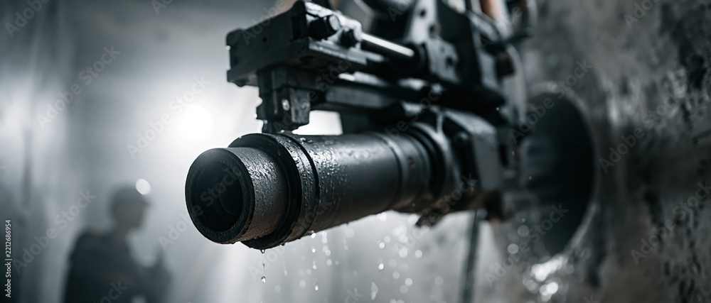 Fototapeta premium A close-up horizontal shot of a powerful wet core drilling machine boring a large hole into a concrete structure with water droplets, featuring a blurred worker in a dark industrial tunnel.