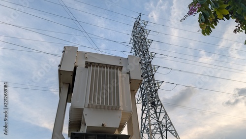 Fotografie High-voltage power transformer installed next to a steel lattice transmission tower under a cloudy sky