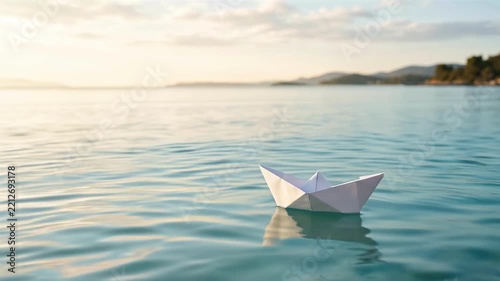 A solitary white paper boat floats gently on the calm, clear blue ocean water with a distant shoreline.