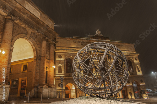 View of the Sferisterio di Macerata with its neoclassical architecture illuminated at night and a large spherical metal sculpture in the foreground in Macerata, Marche, Italy.