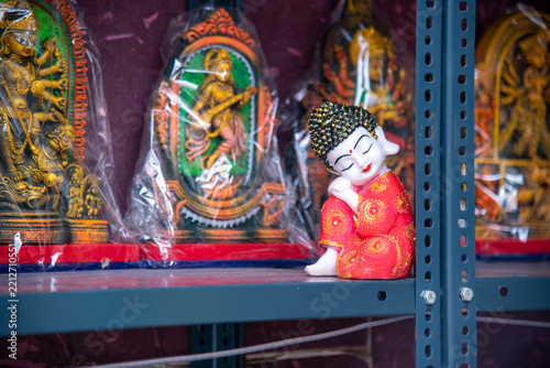 A colorful collection of traditional religious idols displayed on a shelf in a store, featuring a small sleeping monk statue in vibrant red attire with a golden bead head, alongside framed Hindu deity