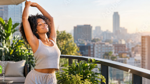 A joyful young Black woman stretching her arms upwards, with her eyes closed and a peaceful smile, embracing the morning sunlight on an urban balcony