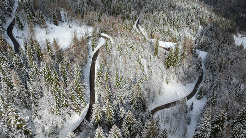 Drone captures majestic snow covered peaks and winding road in Triglav National Park. Traditional wooden cabin sits surrounded by frozen alpine pine forest near Vrsic Pass during winter day. 