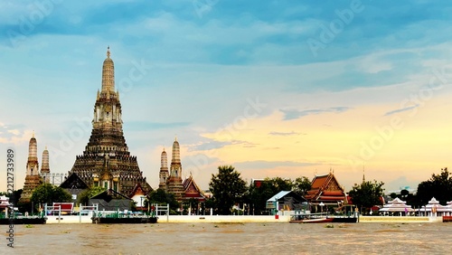 Wat Arun temple rises beside the Chao Phraya River in Bangkok, Thailand, as a long-tail boat cruises past during a peaceful evening with soft clouds and warm sunset light over the riverside.