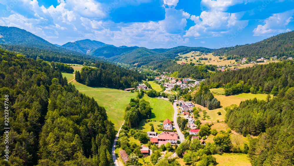 Obraz premium A village road crosses farmland in a wooded Austrian valley.. Red roofs line a curving road among green and yellow fields beneath scattered cloud shadows.