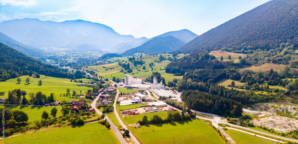 Obraz premium An industrial site stands beside a road in a mountain valley.. Gray towers and pale roofs stand beside a road among green fields beneath hazy ridges.