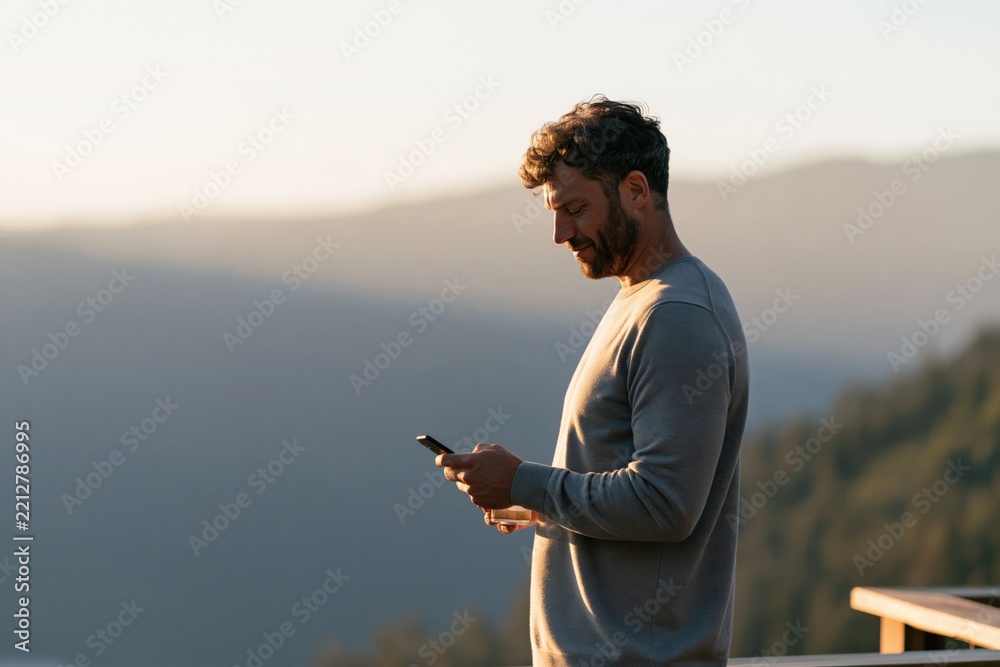 Fototapeta premium Man checking smartphone on balcony at sunset with mountain view for travel lifestyle background and mobile communication banner
