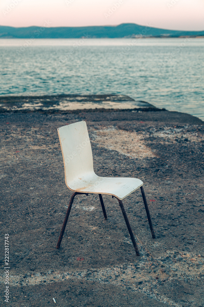 Fototapeta premium A single white chair standing alone on a concrete seaside pier at dusk with calm sea waters in the background.