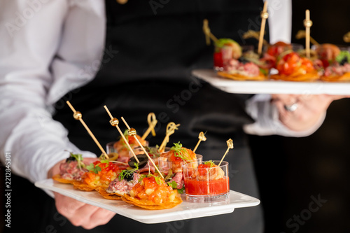 A waiter in a white shirt and black apron holds plates of elegant catering appetizers, including tartlets and shrimp cocktails on skewers, at an event.