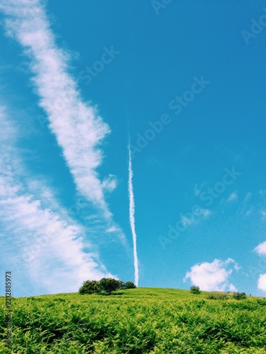 Vertical vapour trail amongst clouds over a lush green hilltop on summer day, UK