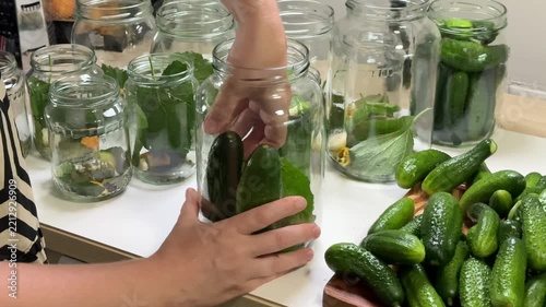 a person places green cucumbers into a glass jar, several other jars containing green vegetables and leaves sit on a white surface, a pile of cucumbers rests on a wooden board