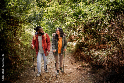A joyful Black couple enjoys a hike on a forest trail, using trekking poles. They share smiles and laughter amidst lush greenery and natural surroundings, creating a vibrant and adventurous atmosphere