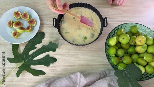 A person uses a pink spatula to stir a pale yellow batter with green fruit pieces in a black fluted baking pan on a light wood surface