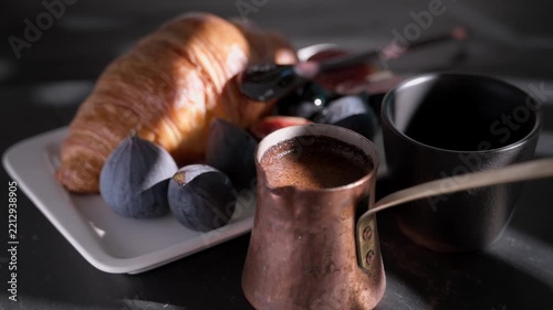 Freshly brewed coffee from a cezve is poured into a cup, breakfast idea croissant and a juicy, ripe fig drizzled with honey, real-time video