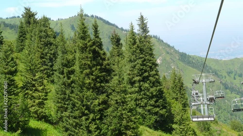 Empty chairlift chairs move up steep mountain slope past tall spruce trees, sunny summer day with hazy valley below, POV ride on ski lift