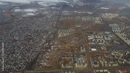 Residential suburbs with private houses, new apartment blocks and straight roads seen from airplane, patches of snow on brown fields, overcast sky, high altitude aerial view