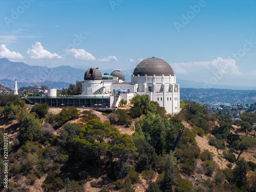 Aerial view captures Griffith Observatory set atop Mount Hollywood with domes, trees, hillside vegetation, and distant mountain ridges in Los Angeles, California on a clear sunny day.