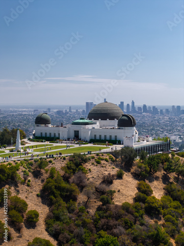 Griffith Observatory sits on the hill above Los Angeles, California, with tourists walking near the historic domes and the distant downtown skyline visible across the horizon.
