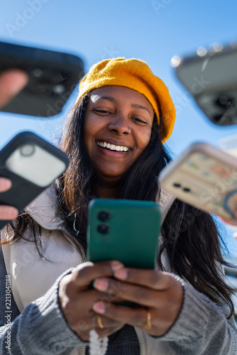 Vertical. Smiling Black woman looks at a smartphone while friends hold phones and cash nearby, illustrating mobile payment, digital finance and shared expenses outdoors.