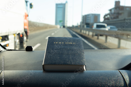 Holy Bible on a car dashboard on Japan's Tomei Expressway, with a bright softly blurred highway and urban scenery symbolizing a peaceful spiritual journey.
