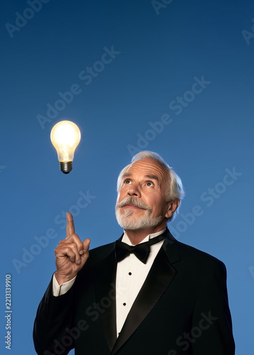 Elderly Gentleman in Formal Tuxedo Pointing Toward Glowing Lightbulb Above Him, Symbolizing Brilliant Idea and Sudden Insight, Blue Background, Vertical Orientation, Copy Space