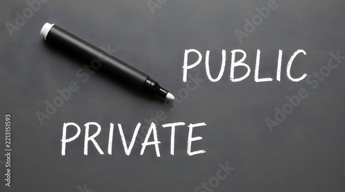 A black marker pen lies diagonally on a dark gray textured surface next to handwritten white text reading public and private, representing legal concepts and business strategy choices clearly