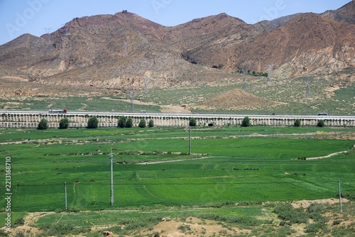 Newly built expressway viaduct crossing green farmland and barley fields beneath arid mountains between Lhasa and Shigatse, Tibet, July 2025.