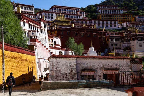 Fotografie A lone Tibetan pilgrim walks through the courtyard of Ganden Monastery in Dagze, Tibet, past traditional whitewashed buildings, golden roofs and a white stupa on a sacred Buddhist hillside