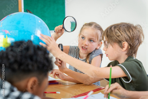 Multiethnic school children using magnifying glass to examine globe in classroom, Diverse group of elementary students studying geography, Curious kids learning science and geography in classroom