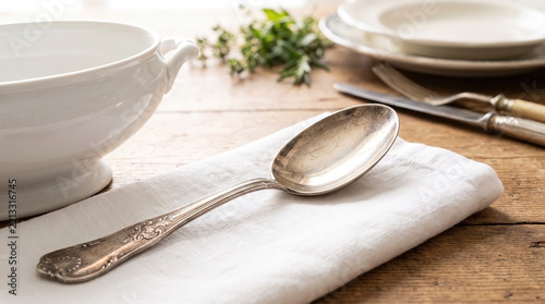 Old restaurant serving spoon on white cloth beside bowl and plate in rustic dining setting. Spoon features ornate design and silver plating,