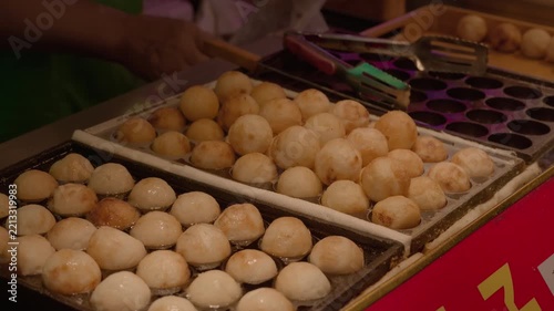 Cooked round savory balls rest on two grilling trays at a street food stall, prepared for serving as a popular snack.
