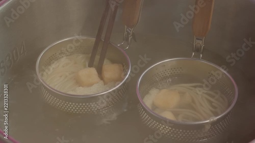 Noodles and other food items are being cooked in boiling hot water using metal strainer baskets with chopsticks.