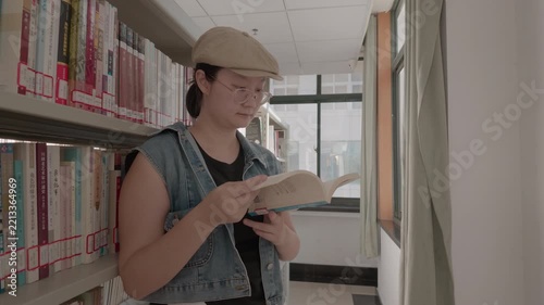 A young adult with glasses reads a book leaning against library bookshelves by a window.