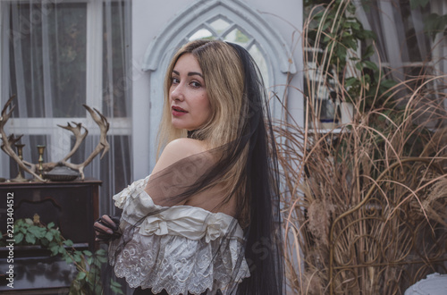 Mysterious gothic bride in a voluminous Victorian lace dress, black velvet corset and dramatic black veil at an ivy-covered old house. Dark romantic Halloween-inspired costume with antique atmosphere