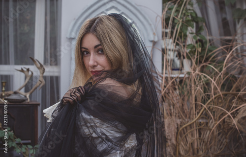 Mysterious gothic bride in a voluminous Victorian lace dress, black velvet corset and dramatic black veil at an ivy-covered old house. Dark romantic Halloween-inspired costume with antique atmosphere