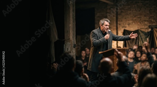 A leader addressing followers from a podium in a dim hall. Dangerous cult gathering and persuasive speech.