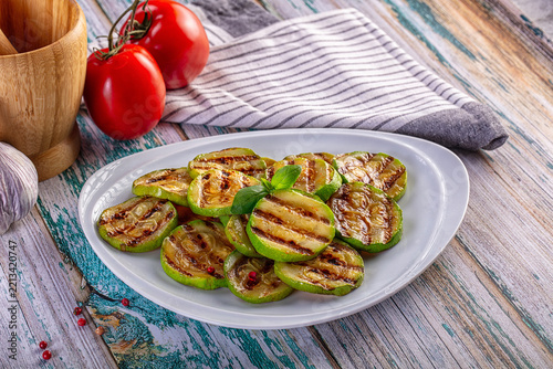 Grilled zucchini slices with basil on a white plate. Healthy vegetarian side dish with fresh tomatoes and garlic on a rustic wooden background