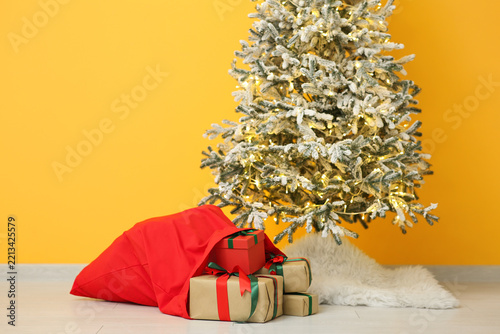 Christmas tree and Santa bag with gift boxes near yellow wall in room