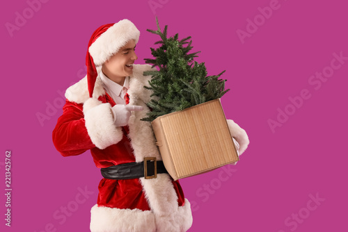 Young Santa Claus pointing at Christmas tree on violet background