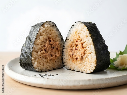Two halves of a tuna onigiri rice ball with nori seaweed wrap, served on a ceramic plate with sesame seeds and ginger garnish, closeup studio shot
