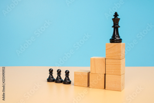 Obraz na plátně Black chess king elevated on wooden block steps above a group of three pawns on a beige surface against a blue background