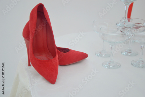 A festive round table with a light tablecloth, on which stand red suede high-heeled shoes, champagne glasses and long red candles with ribbons.