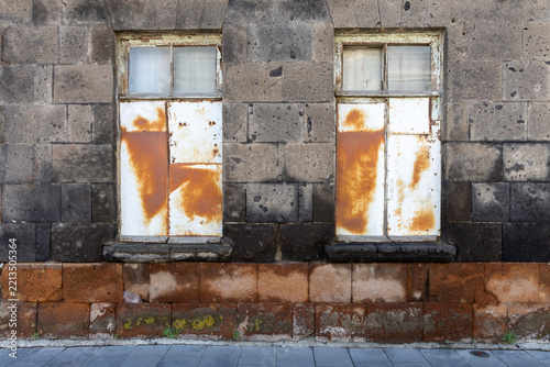 Boarded up windows covered with rusted metal sheets mark the weathered facade of an abandoned black stone building showing signs of urban decay, Gyumri, Armenia