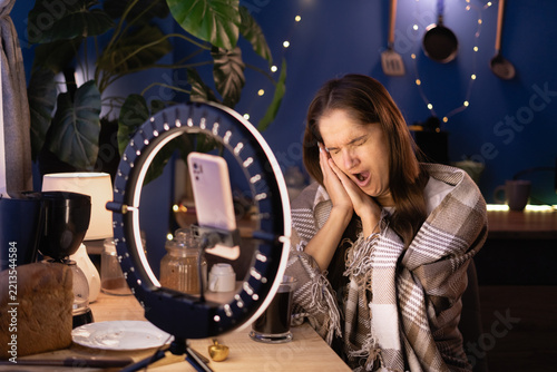 Woman influencer covering warm blanket holding hands in sleep gesture yawning sleepy films video content for social media using phone with ring light at home, live streaming.