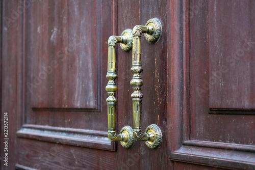 Pair of vintage brass door handles on heavy dark brown wooden double panel entrance door indoors. Classic antique bronze pull hardware accessory, retro luxury interior architecture decoration element.